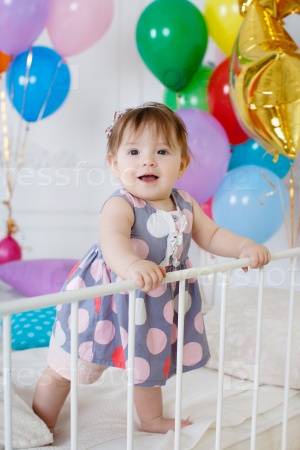Little girl with red hair and dark eyes, wearing a gray dress with white and pink polka dots, sleeveless, standing on a white bed in the background of colorful balloons and gold stars on his first birthday