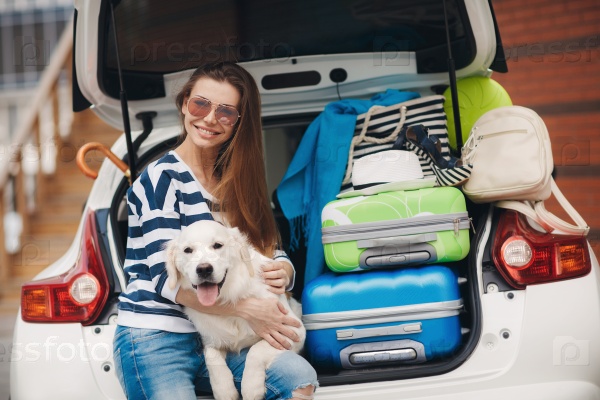 Beautiful young woman brunette with long hair, a beautiful smile, wearing sun glasses, dressed in a t-shirt with white and dark blue stripes, blue jeans with holes in the knees, sits with his white dog near a white car loaded with stuff