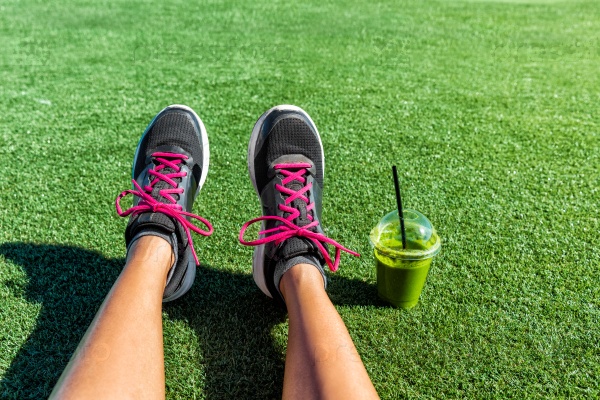 Healthy lifestyle runner girl running shoes selfie with green smoothie. Fitness woman drinking juice to go after workout in park. Healthy lifestyle sporty female athlete POV mobile phone feet picture.