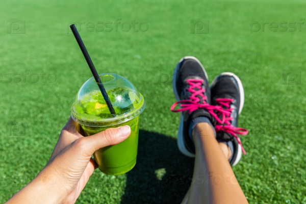 Green smoothie woman drinking plastic cup breakfast takeaway juice to go after morning run in summer park. Healthy lifestyle sporty person POV of hand holding drink with running shoes feet selfie.