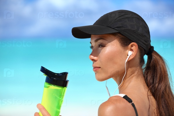 Healthy sporty Asian runner woman running on beach drinking water bottle listening to music with earbuds and smartphone wearing sun cap for solar protection during summer. Active living.