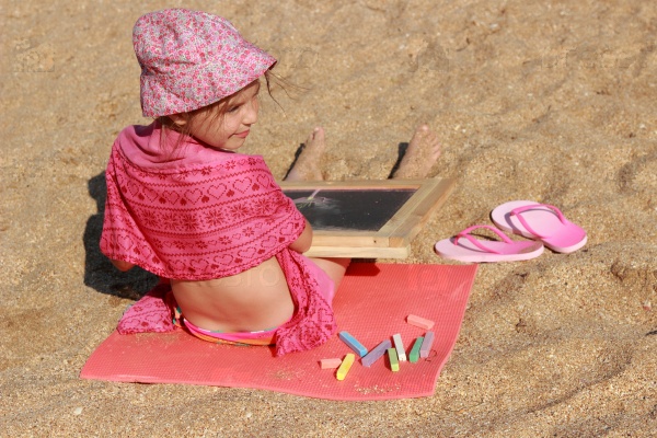 Charming little girl sitting on the beach in the sand, and draws on the board