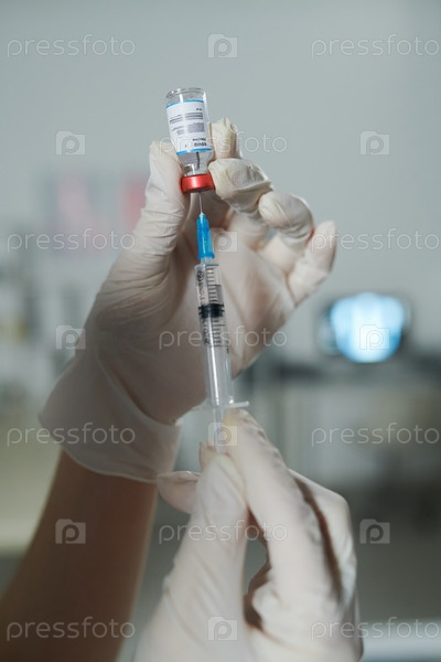 Gloved hands of clinician or nurse holding syringe and small bottle with new vaccine