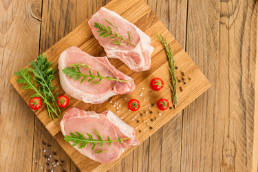Три идеальных куска свежей свинины на разделочной кухонной доске с листьями рукколы и помидорами. вид сверху - MarinaMos