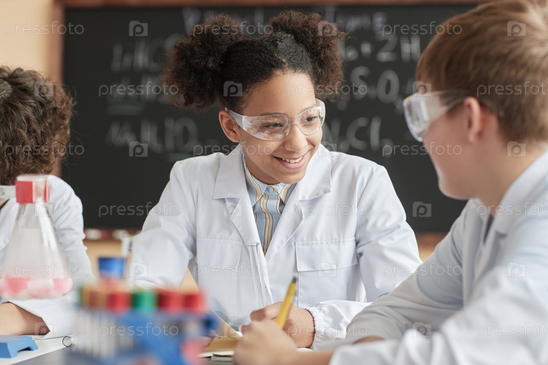 Smiling black schoolgirl wearing lab coat in science class talking to friend.