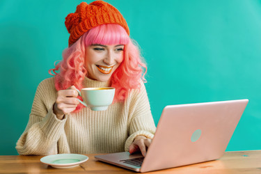 Happy girl with pink hair enjoys coffee while working on her laptop in a cozy café. Generative AI - RomanZ