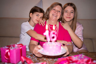 Женщина празднует 16-летие с двумя дочерьми, розовый торт со свечами, яркие подарки и праздничные украшения в уютной обстановке внутри помещения - Darya Petrenko