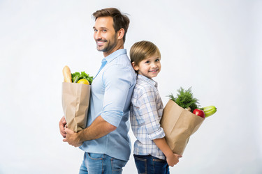Отец и сын улыбаются, держа в руках свежие продукты в бумажных пакетах на белом фоне - RomanZ