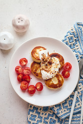 Вкусные сырники (оладьи с сыром), политые сметаной, посыпанные хрустящим жареным луком и свежими помидорами черри, подаются на белой тарелке, накрытой синей вязаной скатертью. Идеально подходят для завтрака или бранча - Irrin0215
