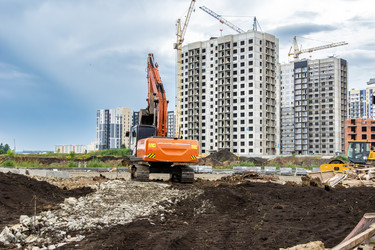 Оранжевый экскаватор и желтый дорожный каток работают на неровной местности с грунтом и щебнем перед бетонными каркасами высотных зданий и башенными кранами в Кемеровской области Кузбасс (выборочная фокусировка) - dkorostilev78