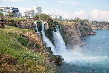 Нижний водопад Дюден низвергается в Средиземное море, на поверхности воды появляется радуга, а на смотровой площадке у скалы сидит фотограф. Летний пейзаж, Анталья, Турция - AlexTelford