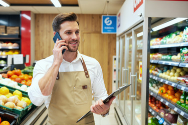 Взрослый мужчина, менеджер продуктового магазина, разговаривает по смартфону в супермаркете. - SeventyFour