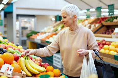 Пожилая женщина выбирает свежие фрукты для здоровых покупок в продуктовом магазине. - SeventyFour