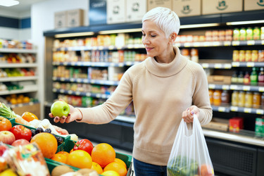 Женщина средних лет осматривает яблоко, выбирая полезный продукт в магазине. - SeventyFour