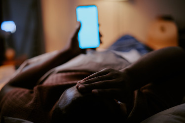 Чернокожий мужчина, лежащий в постели и держащий в руках смартфон из-за нарушения сна - SeventyFour