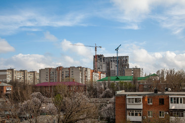 Городской пейзаж с многоэтажными жилыми домами, двумя действующими строительными кранами, распускающимися деревьями под ярко-голубым небом с рассеянными облаками. Теплый солнечный свет подчеркивает кирпичные фасады и зеленые крыши - Kathleen77