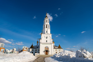 Екатеринбург, Россия. Белый православный храм с золотыми куполами, религиозные фрески над входом, снежные заносы и современные здания под голубым небом - NikitaMaykov