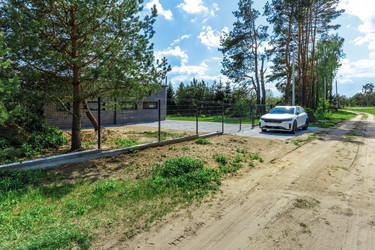 Внешний вид современного загородного дома с надежным металлическим забором и мощеной подъездной дорожкой, где припаркован белый автомобиль. Участок окружен соснами и зеленой природой, расположен рядом с сельской грунтовой дорогой под ярким солнечным небом - deek
