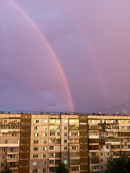 Многоэтажное здание с окнами, освещенное теплым светом. На фоне — радуга и фиолетовое небо - Blic