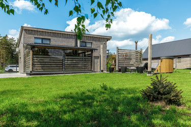 Живописный вид на задний двор современного загородного дома с крытой террасой, оборудованной защитными шторами, и открытой спа-зоной с гидромассажными ваннами. На переднем плане — аккуратный зеленый газон, обрамленный березовыми ветвями и небольшим хвойны - deek