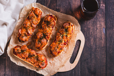 На столе лежат булочки для хот-догов с сыром, сосисками, помидорами и зеленью, разложенные на бумаге - Elena Rui
