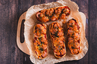 Крупный план испеченных булочек для хот-догов с сыром, сосисками, помидорами и зеленью, разложенных на бумаге на столе - Elena Rui