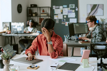 Чернокожая женщина закрывает лицо во время работы в напряженном офисе - AnnaStills