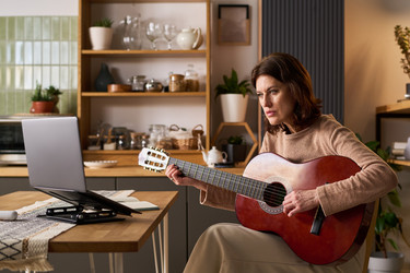 Женщина средних лет практикуется на гитаре во время виртуального урока дома - Pressmaster