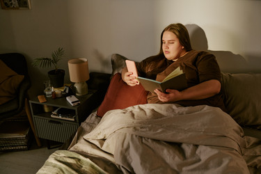 Женщина больших размеров читает книгу и пользуется смартфоном ночью в постели - AnnaStills