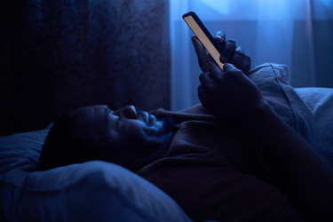 Чернокожий взрослый мужчина просматривает смартфон в постели из-за нарушения сна - SeventyFour