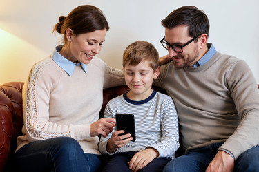 Счастливые родители и сын вместе пользуются смартфоном на диване - DragonImagesAsia