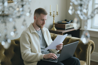 Сосредоточенный мужчина, работающий за ноутбуком и читающий документы на диване - DragonImagesAsia