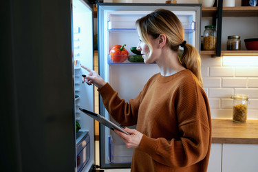 Женщина с цифровым планшетом в руках проверяет продукты в открытом холодильнике - DragonImagesAsia