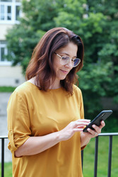 Улыбающаяся женщина в очках пользуется смартфоном на открытом воздухе - DragonImagesAsia