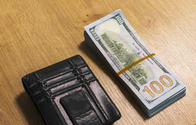 На деревянном столе рядом с черным кожаным кошельком лежит пачка стодолларовых купюр. Деньги - Pazemin