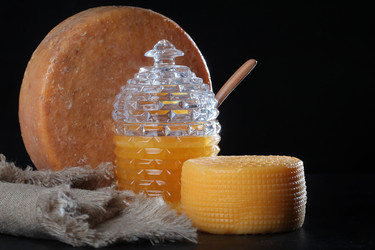 Натюрморт с премиальными медовыми и сырными продуктами. Хрустальный горшок для меда в форме улья. Золотисто-желтый мед в стеклянной банке. Деревянная ложка для меда помещена внутрь горшка. Сзади стоит большой круглый кусок выдержанного сыра. Справа лежит  - RusAKphoto
