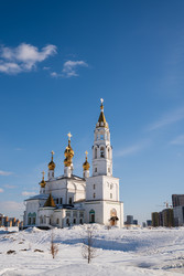 Екатеринбург, Россия. Белый православный храм с золотыми куполами и колокольней, окруженный заснеженной землей и современными высотными зданиями под ясным голубым небом - NikitaMaykov