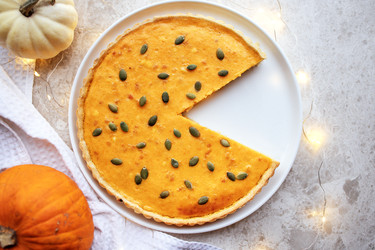 Тыквенный пирог с отрезанным кусочком, украшенный тыквенными семечками, выставлен на белой тарелке в окружении декоративных тыкв и мягкого освещения, создавая теплую осеннюю атмосферу - Darya Petrenko