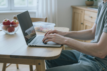 Мужчина печатает на ноутбуке за кухонным столом, работая из дома - DragonImagesAsia