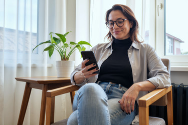 Улыбающаяся женщина, сидящая в кресле и пользующаяся смартфоном дома - DragonImagesAsia