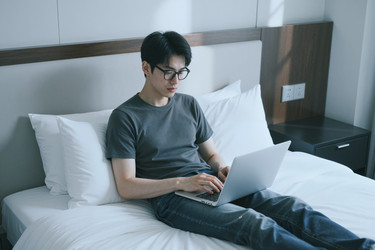 Азиат работает за ноутбуком в постели во время удаленной работы - DragonImagesAsia