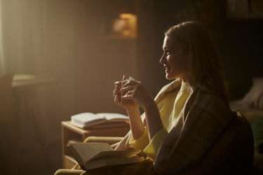 Улыбающаяся женщина с горячим напитком в руках и читающей книгой в уютной комнате - DragonImagesAsia