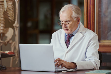 Пожилой врач-мужчина работает за ноутбуком в медицинском кабинете - DragonImagesAsia