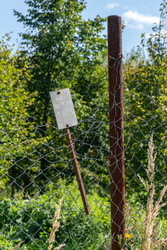 A close‑up of an old metal sign without any inscriptions, behind a chain‑link fence, against a backdrop of green trees and a blue sky on a sunny summer day - DmitryKurullin