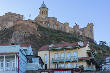 Вид снизу на церковь Сурб Геворг (церковь Святого Георгия), старинные дома исторического района Тбилиси Кала и крепостной комплекс Нарикала на фоне голубого неба, Грузия - gumbao