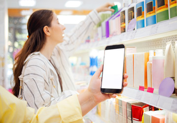покупательница держит в руках смартфон с пустым экраном, выбирая косметические средства с полок в современном розничном магазине с видимой красочной упаковкой - Darya Petrenko