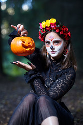 Цщьфт with Santa Muerte make-up sits on a path in forest and holds a Halloween pumpkin by the stalk - Serg Zastavkin