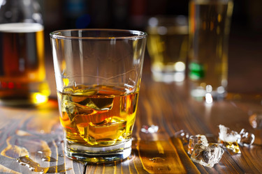 Glass of alcohol with ice cubes sits on a bar top with bottles of liquor in the background - yliv