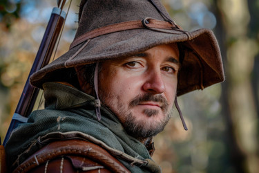 Archer wearing leather hat standing in forest holding bow - yliv