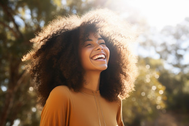 Carefree young woman with curly hair laughing with joy under the sunlight in nature - yliv